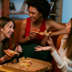 three young women sharing pizza and laughing in an apartment